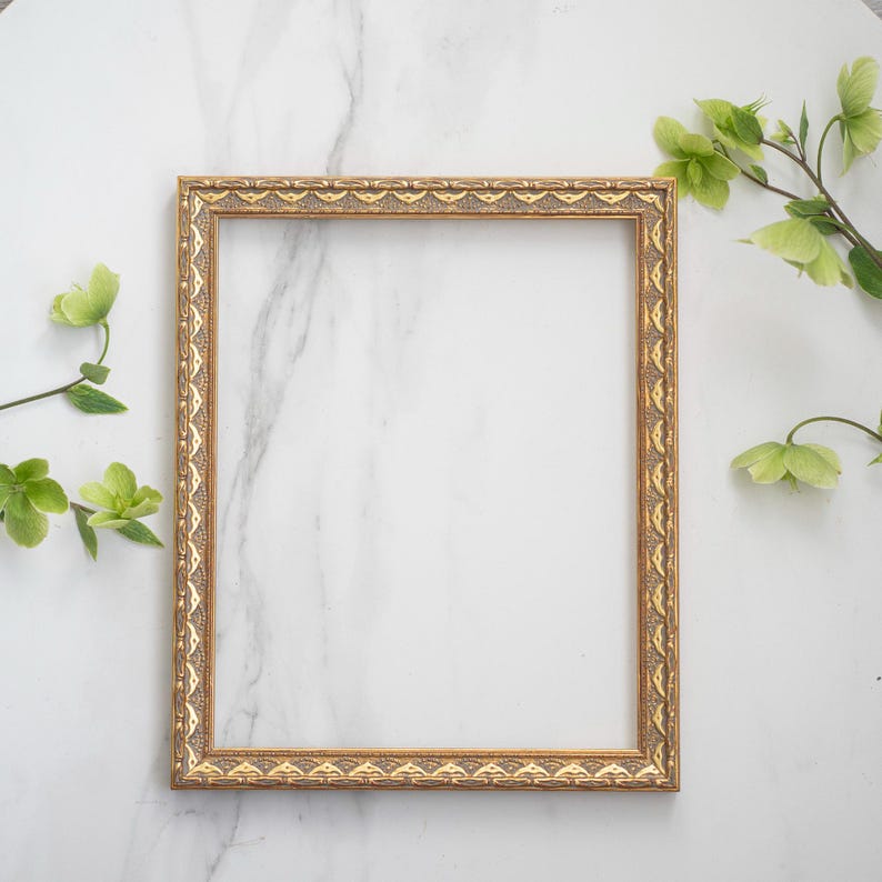 May include: A gold picture frame with an ornate design sits on a white marble surface. Green leaves and flowers are arranged around the frame.