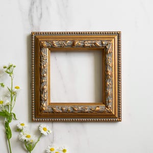 Könnte beinhalten: Quadratischer goldfarbener Bilderrahmen mit aufwendigen Details und einem Perlenrand. Der Rahmen ist leer, vor einem weißen Marmorhintergrund. Weiße Gänseblümchen mit gelben Zentren befinden sich unten links.