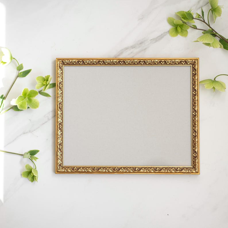 May include: A gold picture frame with an ornate design. The frame is on a white marble surface.