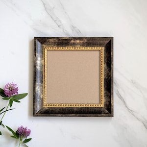 May include: A square picture frame featuring a wide, distressed black and gold outer border and a thinner, ornate gold inner trim. The center of the frame contains a plain, light tan mat board, providing a neutral space for display.