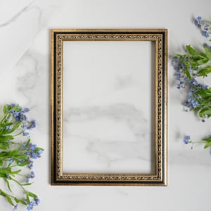 Peut inclure: Un cadre photo doré avec une bordure intérieure noire est posé sur une surface en marbre blanc. Le cadre est vide et prêt pour une photo ou une œuvre d'art. Des fleurs bleues de myosotis se trouvent de chaque côté du cadre.