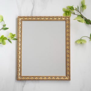 May include: A gold picture frame with an ornate design sits on a white marble surface. Green leaves and flowers are arranged around the frame.
