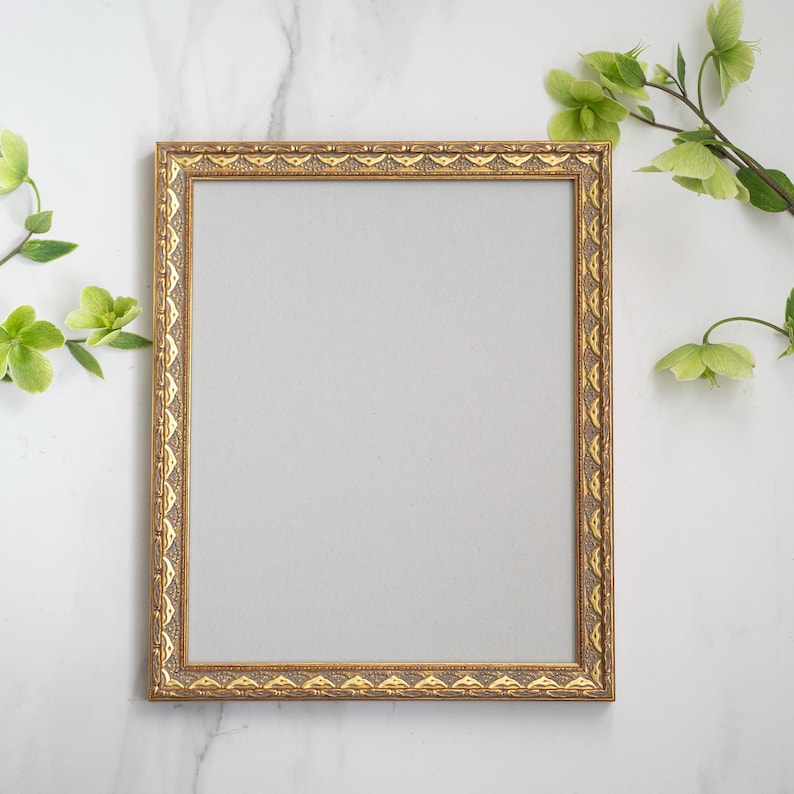 May include: A gold picture frame with an ornate design sits on a white marble surface. Green leaves and flowers are arranged around the frame.
