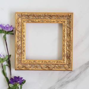 Op de afbeelding: Een vierkante fotolijst met een metallic gouden afwerking en ingewikkelde reliëf bloemendetails langs de rand. Naast de lijst staat een slanke groene stengel met twee paarse asterbloemen met gele harten en groene bladeren.