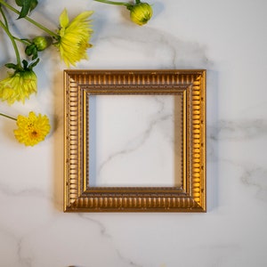 May include: A square picture frame with a metallic gold finish and a textured, repeating vertical groove pattern along its border. The frame features a stepped profile with intricate detailing and a dark, recessed inner edge.