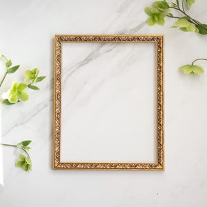May include: A gold-colored ornate picture frame with a white background. The frame is on a white marble surface.