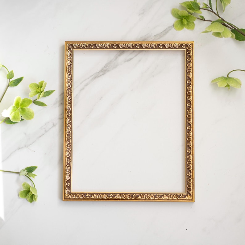 May include: A gold-colored ornate picture frame with a white background. The frame is on a white marble surface.