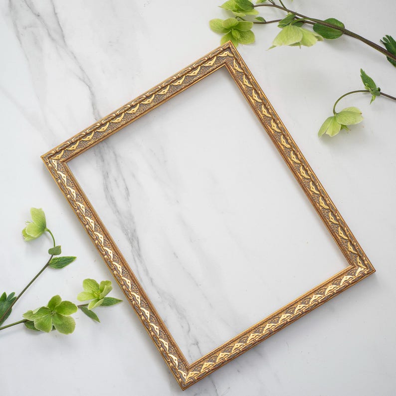 May include: A gold picture frame with an ornate design sits on a white marble surface. Green leaves and flowers are arranged around the frame.