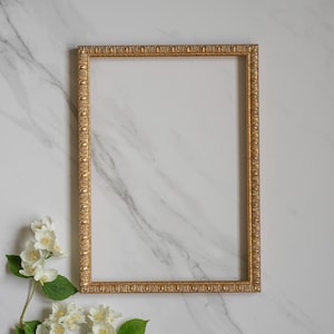 May include: A rectangular picture frame with a metallic gold finish and an ornate, repeating embossed pattern along the edges. The frame is empty, featuring a clean, hollow center, and is positioned vertically against a white marble surface.