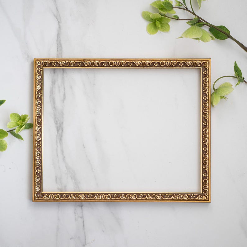 May include: A gold-colored picture frame with an ornate design sits on a white marble surface. Green leaves and flowers are arranged around the frame.
