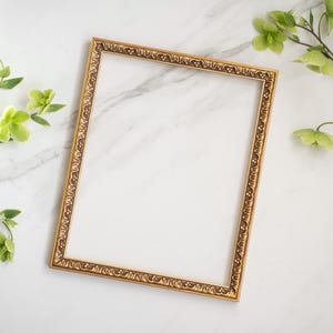 May include: A gold-colored ornate picture frame with a white background. The frame has a decorative design around the edges.