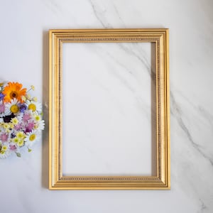 May include: A gold-colored picture frame with a decorative border sits on a white marble surface.