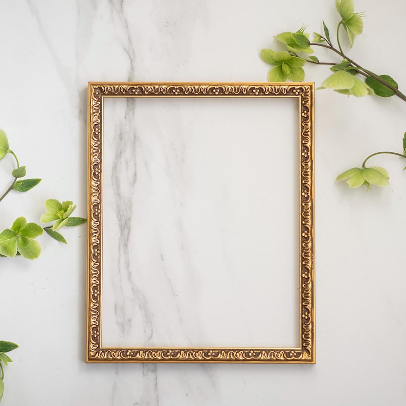 May include: A gold ornate picture frame on a white marble background. The frame is empty and ready for a photo or artwork.