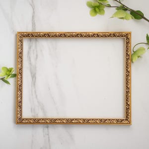 May include: A gold-colored picture frame with an ornate design sits on a white marble surface. Green leaves are visible in the background.