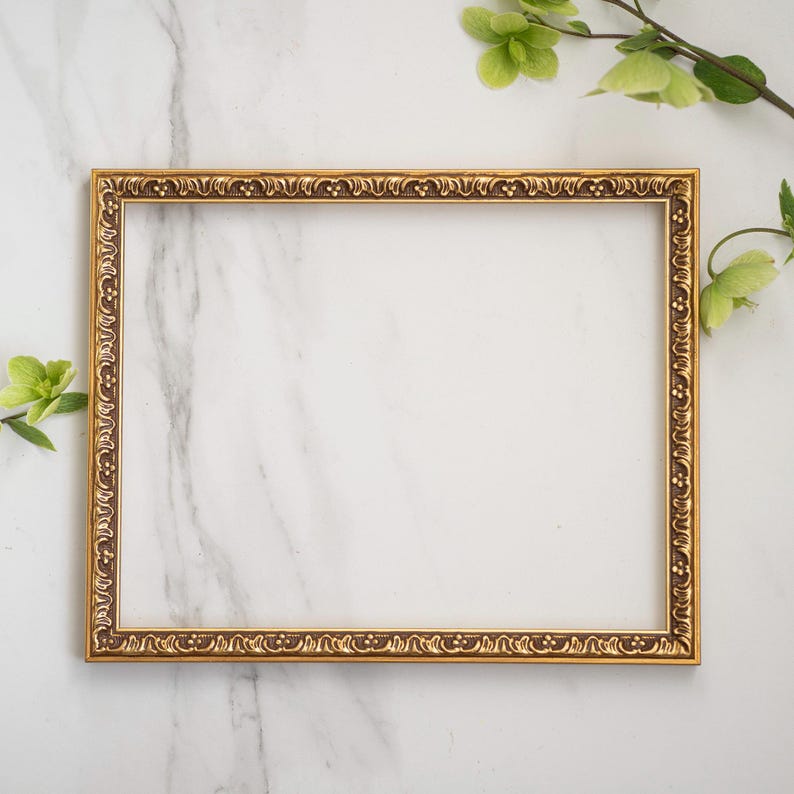 May include: A gold-colored picture frame with an ornate design sits on a white marble surface. Green leaves are visible in the background.