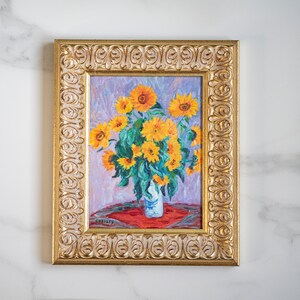 May include: A framed painting of a bouquet of sunflowers in a vase. The sunflowers are yellow and the vase is white. The painting is set against a light purple background. The frame is gold and has a decorative design.