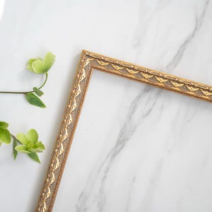 May include: A gold picture frame with an ornate design sits on a white marble surface. Green leaves and flowers are visible in the background.