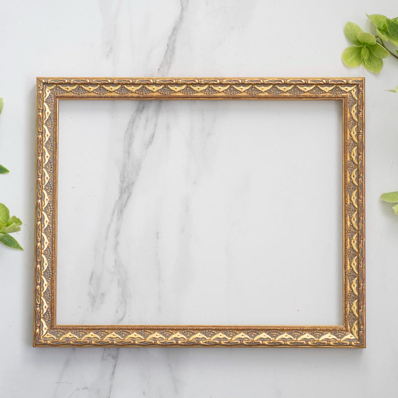 May include: A gold picture frame with an ornate design sits on a white marble surface. The frame is empty and ready for a photo or artwork.