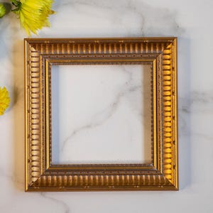Könnte beinhalten: Ein rechteckiger Bilderrahmen mit metallisch goldener Oberfläche, der ein sich wiederholendes vertikales Rillenmuster entlang der Innen- und Außenkanten sowie einen glatten, vertieften Mittelrand aufweist. Der Rahmen ist leer und strukturiert.