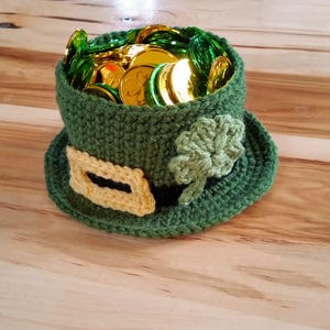 Może przedstawiać: Zielony, szydełkowy kapelusz leprechauna z złotą klamrą i wzorem koniczyny. Kapelusz jest wypełniony złotymi monetami.