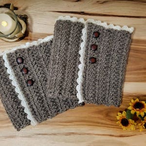 Puede incluir: Dos calentadores de piernas de color marrón hechos a crochet con ribete de encaje blanco y botones marrones. Los calentadores de piernas están sobre una superficie de madera.