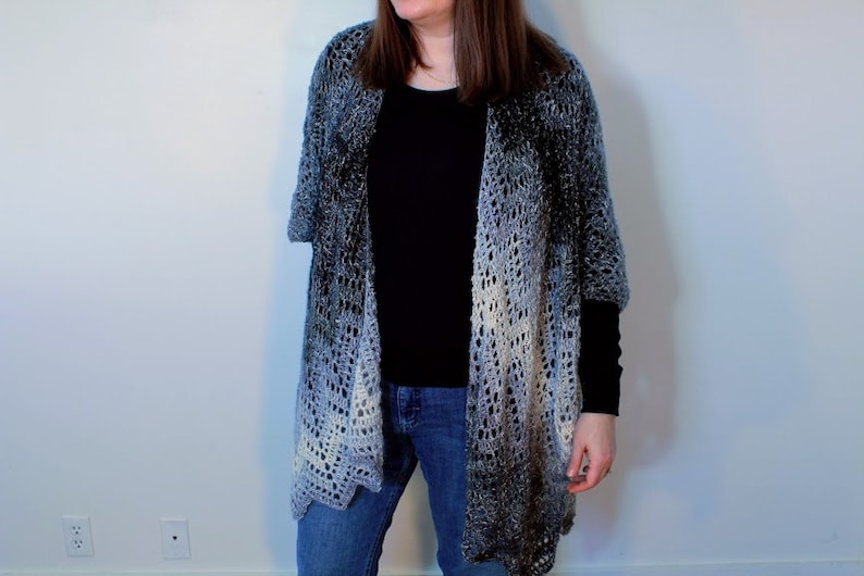 May include: A gray and white ombre crochet shawl with a lacy pattern. The shawl is draped over a person wearing a black shirt and blue jeans.