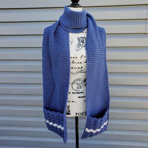 May include: A blue crochet scarf with a built-in cowl and pockets. The scarf is displayed on a mannequin. The pockets have a white wavy trim. The scarf is made of a textured yarn.