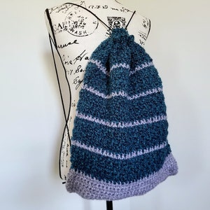 Puede incluir: Una bolsa de cordón de ganchillo de color verde azulado y morado con un patrón de rayas. La bolsa está colgada de un maniquí blanco.