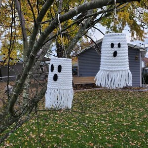 May include: Two white crocheted ghost decorations with black eyes and mouths hanging from a tree branch. The ghosts have fringe at the bottom.