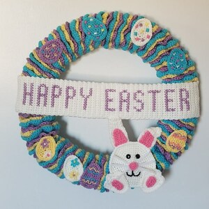 May include: A crocheted Easter wreath with a purple, yellow, and blue color scheme. The wreath is decorated with crocheted eggs and a bunny. The wreath has a white banner that says "Happy Easter".