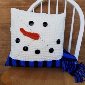 May include: White crocheted cushion with a snowman face and black buttons for eyes and a mouth. The cushion has a blue and white striped trim and a blue crocheted scarf with fringe.