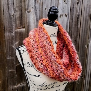 Könnte beinhalten: Ein gehäkelter Infinity-Schal in Orange- und Rosatönen. Der Schal wird auf einer weißen Schaufensterpuppe mit schwarzer Schrift präsentiert.