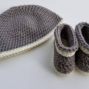 May include: Gray crocheted baby hat and booties with a white trim. The hat is slouchy and the booties have a pointed toe.