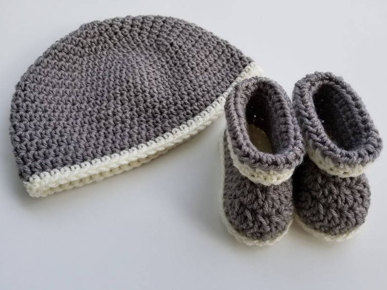 May include: Gray crocheted baby hat and booties with a white trim. The hat is slouchy and the booties have a pointed toe.
