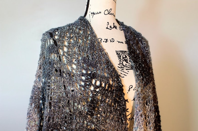 May include: A gray and white crocheted cardigan with a loose, open weave. The cardigan is draped over a dress form.