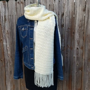 Peut inclure: Une écharpe en crochet de couleur jaune pâle avec des franges aux extrémités. L'écharpe est drapé sur une veste en jean.