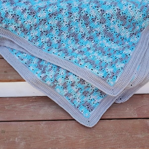 Könnte beinhalten: Eine gehäkelte Babydecke in Blautönen und Grau mit einem strukturierten Muster. Die Decke ist gefaltet und über eine Holzoberfläche gelegt.