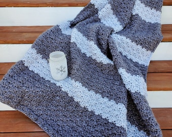 Pode incluir: Uma manta de crochê listrada em cinza e branco com um padrão texturizado. A manta está dobrada e drapeada sobre uma escada de madeira.
