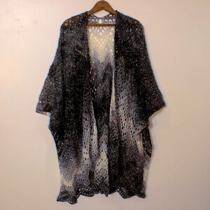 May include: A black and white ombre crochet shawl with a lacy pattern. The shawl is hanging on a wooden hanger against a white wall.