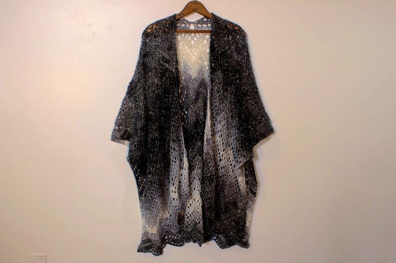 May include: A black and white ombre crochet shawl with a lacy pattern. The shawl is hanging on a wooden hanger against a white wall.