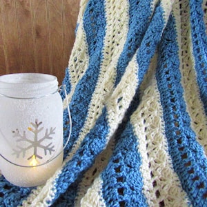 May include: A white crocheted blanket with blue stripes. The blanket is draped over a frosted glass jar with a snowflake design and a lit candle inside.