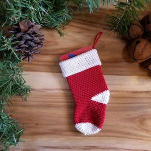 May include: A red and white crocheted Christmas stocking with a white cuff and a white toe. The stocking is filled with a gift card.