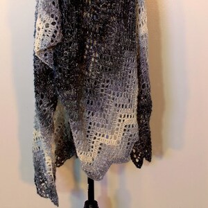 May include: A crocheted shawl in shades of gray and white with a lacy pattern. The shawl is hanging on a black stand against a white wall.