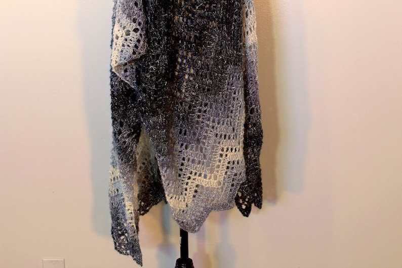 May include: A crocheted shawl in shades of gray and white with a lacy pattern. The shawl is hanging on a black stand against a white wall.