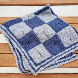 Pode incluir: Uma manta de crochê com um padrão xadrez em tons de azul e cinza. A manta tem uma borda dupla em um cinza mais claro.