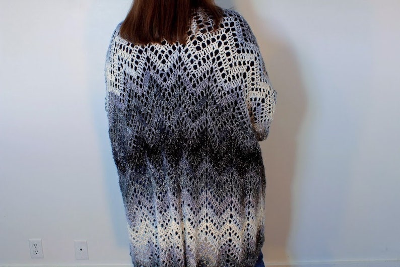 May include: A long, crocheted cardigan in a gradient of white, gray, and black. The cardigan has a loose fit and a delicate openwork pattern.