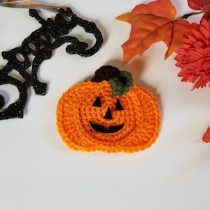 May include: A crocheted orange pumpkin with a black jack-o'-lantern face. The pumpkin has a green stem and is surrounded by fall leaves.