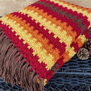 May include: A crocheted throw blanket with a striped pattern in shades of red, orange, yellow, and brown. The blanket has a fringe border.