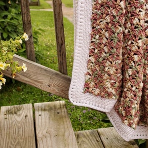 May include: A crocheted throw blanket with a pink, green, and cream color pattern. The blanket has a white crocheted border and is draped over a wooden railing.