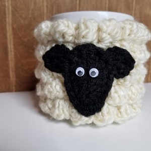 May include: White crocheted mug cozy shaped like a sheep with a black wool face and white googly eyes.